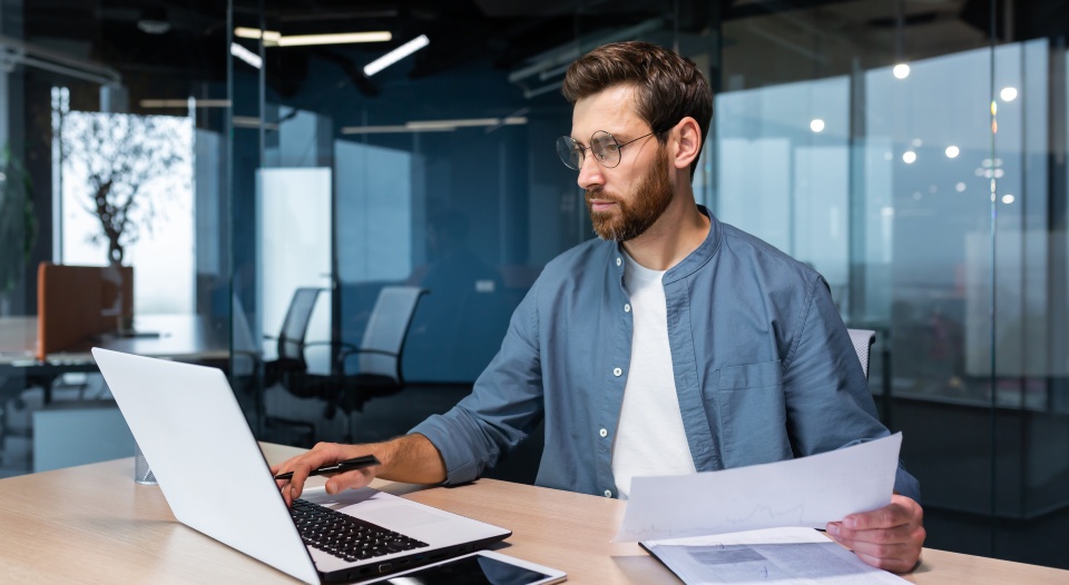 Serious and focused financier accountant on paper work inside office, mature man using calculator and laptop for calculating reports and summarizing accounts, businessman at work in casual clothes.