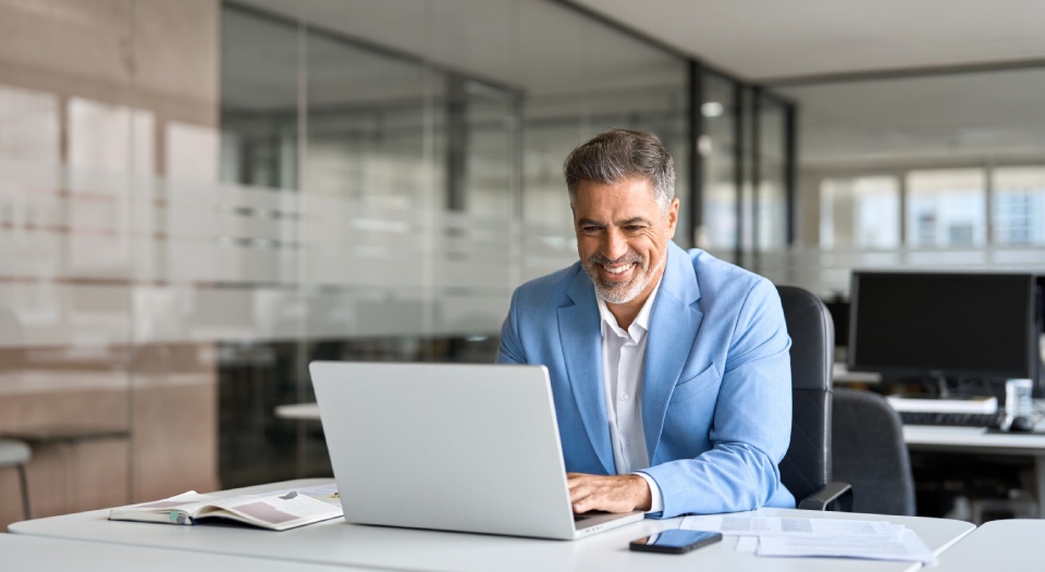 Happy senior 50 years old professional business man wearing suit using computer sitting at desk. Busy smiling middle aged businessman executive investor bank manager working on computer in office.
